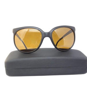 May include: Black sunglasses with amber-colored lenses rest on a black rectangular case. The sunglasses have a rounded frame and a classic design. The lenses are a warm, golden hue, and the case appears to be made of a matte material.
