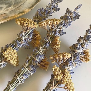 May include: Three bundles of dried lavender and yellow yarrow flowers tied together with thin white string. The lavender stems are purple and blue, while the yarrow clusters are a muted golden yellow. The bundles are arranged in a cluster.