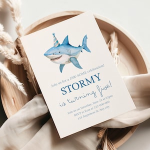 Puede incluir: Una invitación de cumpleaños con una ilustración en acuarela de un tiburón azul sonriente con un gorro de fiesta. La invitación dice "¡STORMY cumple cinco años!" en escritura azul, con detalles del evento debajo. La invitación está en una tarjeta blanca.