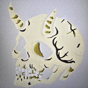May include: Embroidered skull design with horns and intricate detailing. The skull is primarily off-white with accents of gold and black, creating a textured appearance. The horns and other details are highlighted with gold.