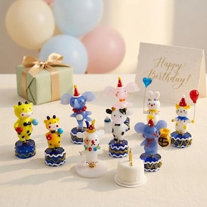 May include: A collection of colorful animal figurines, each wearing a party hat and standing on a blue and gold base. A small white cake with a single candle is in the foreground, with a gift box and balloons in the background. A card reads "Happy Birthday!"