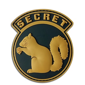 May include: A round patch with a gold squirrel design on a dark teal background. The word "SECRET" is arched across the top in gold lettering. The patch has a gold border.