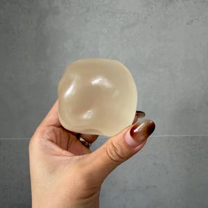 May include: A translucent, beige-colored, rounded object is held in a hand. The object has a smooth, slightly irregular surface. The background is a plain, light gray wall.
