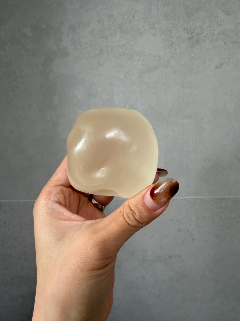 May include: A translucent, beige-colored, rounded object is held in a hand. The object has a smooth, slightly irregular surface. The background is a plain, light gray wall.