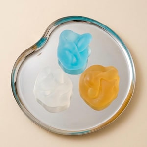 Puede incluir: Una bandeja plateada en forma de riñón contiene tres esculturas abstractas de forma orgánica. Una es azul claro, otra blanca y la otra amarilla. Las esculturas tienen un aspecto liso y translúcido sobre un fondo neutro.