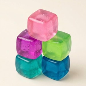 May include: A stack of five translucent, square-shaped ice cubes in vibrant colors. The top cube is pink, with a purple cube and a bright green cube below it, followed by a teal cube and a dark blue cube at the base. Each cube has a smooth finish.