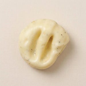 May include: A close-up shot of a light yellow, irregularly shaped soap bar with three deep grooves. The soap has small, dark specks throughout, and the surface appears smooth and slightly glossy. The background is a plain, light beige fabric.