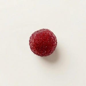 May include: A single, round, red fruit with a textured surface, possibly a waxberry or similar berry, is centered against a plain white background. The fruit's color is a deep, saturated red, with a slightly granular appearance.