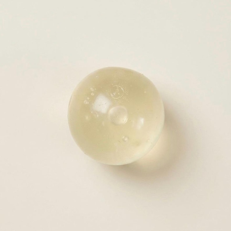 May include: A translucent, spherical object with a slightly yellowish hue. The sphere has a smooth surface with small, internal bubbles and a central, circular indentation. The object is set against a plain, off-white background.