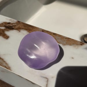 May include: A smooth, lavender-colored soap bar with an organic, rounded shape. The soap is sitting on a marble countertop with brown and white veining. The lighting creates highlights and shadows on the soap's surface.