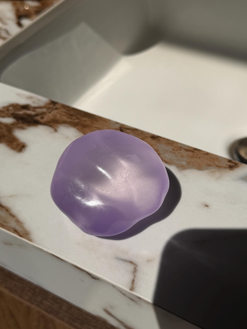 May include: A smooth, lavender-colored soap bar with an organic, rounded shape. The soap is sitting on a marble countertop with brown and white veining. The lighting creates highlights and shadows on the soap's surface.