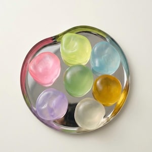May include: A decorative silver-colored dish with a wavy rim holds seven colorful, smooth, rounded objects. The objects are in shades of pink, green, yellow, blue, purple, and gold. The dish has a clear, colorful rim.