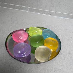 May include: A collection of colorful, smooth, gel-like stones arranged on a silver, circular tray. The stones are in shades of pink, green, blue, yellow, and purple. The tray reflects the colors of the stones.