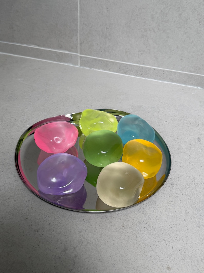 May include: A collection of colorful, smooth, gel-like stones arranged on a silver, circular tray. The stones are in shades of pink, green, blue, yellow, and purple. The tray reflects the colors of the stones.