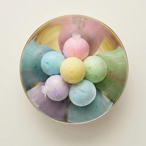 May include: A circular metal bowl containing six pastel-colored spherical bath bombs arranged in a flower pattern. The bath bombs are pink, light blue, yellow, light green, lavender, and teal, each featuring a small clear plastic wrapper tie.
