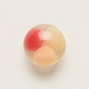 Op de afbeelding: Een glad, bolvormig object van helder materiaal met drie afzonderlijke, afgeronde interne vormen. Eén vorm is felrood, een andere is lichtgeel en de derde heeft een zachte perzikkleur, allemaal ingesloten in de transparante bol.