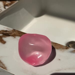 May include: A translucent, pink, heart-shaped soap bar rests on a marble-patterned surface. The soap has a smooth, rounded form with a darker pink hue at its base, reflecting light. The background includes a white sink.