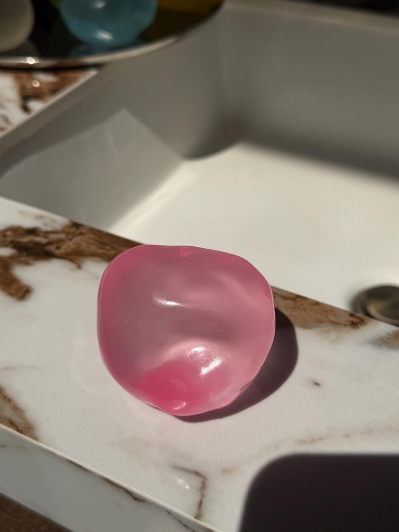May include: A translucent, pink, heart-shaped soap bar rests on a marble-patterned surface. The soap has a smooth, rounded form with a darker pink hue at its base, reflecting light. The background includes a white sink.