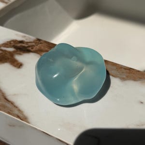 May include: A translucent, light blue, organic-shaped object. The object has a smooth, glossy surface and appears to be made of a solid material. It sits on a white and brown marble-like surface, with a white background.