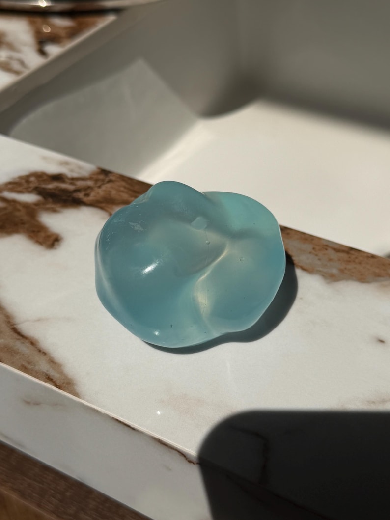 May include: A translucent, light blue, organic-shaped object. The object has a smooth, glossy surface and appears to be made of a solid material. It sits on a white and brown marble-like surface, with a white background.