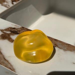 May include: A translucent, yellow, irregularly shaped object, possibly a decorative soap or a bath bomb, rests on a marble-patterned surface. The object's smooth surface reflects light, creating highlights and shadows.