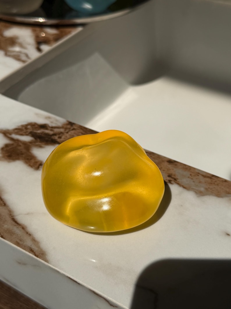 May include: A translucent, yellow, irregularly shaped object, possibly a decorative soap or a bath bomb, rests on a marble-patterned surface. The object's smooth surface reflects light, creating highlights and shadows.