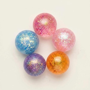 Può includere: Cinque sfere colorate piene di glitter disposte a cerchio. Le sfere sono nelle tonalità di rosa, blu, viola e arancione, con scintillanti accenti glitterati. Lo sfondo è bianco.