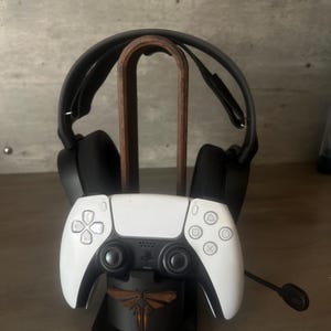 Op de afbeelding: Een gamingheadset en een wit-zwarte controller worden weergegeven op een zwarte standaard. De standaard heeft een houten boog voor de headset en een basis met een vuurvliegontwerp. De controller heeft een PlayStation-logo.