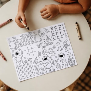 May include: A black and white birthday activity placemat with the text "JORDAN'S BIRTHDAY" and Sesame Street characters. Includes a maze, tic-tac-toe, and a word search. Crayons are on the table.