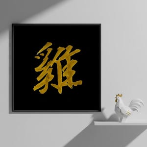 Puede incluir: Una impresión artística cuadrada negra enmarcada con caligrafía china dorada. La obra de arte se exhibe en una pared blanca, con una pequeña figura de gallo blanco en un estante debajo. La caligrafía es el punto focal.