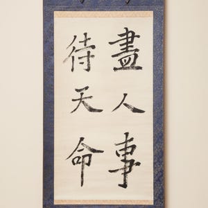 May include: A traditional Japanese scroll with calligraphy in black ink on cream-colored paper. The scroll is framed by a blue fabric with a floral pattern and wooden dowels at the top and bottom. The text is in Japanese.