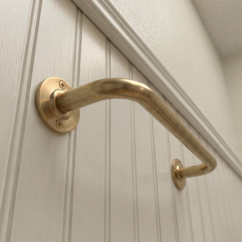 May include: A curved brass grab bar mounted on a white paneled wall. The bar has a rounded shape and is attached to the wall with circular mounting plates secured by screws. The design is simple and functional.