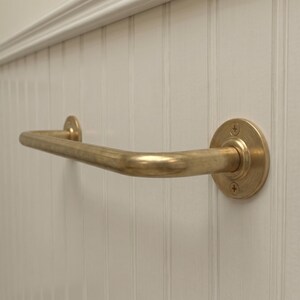 May include: A brass-colored grab bar with a curved design, mounted on a white, vertically paneled wall. The bar has a circular mounting plate on one end and a smaller, matching plate on the other.