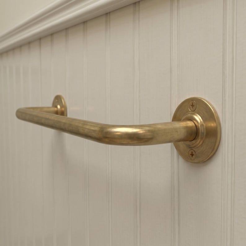 May include: A brass-colored grab bar with a curved design, mounted on a white, vertically paneled wall. The bar has a circular mounting plate on one end and a smaller, matching plate on the other.