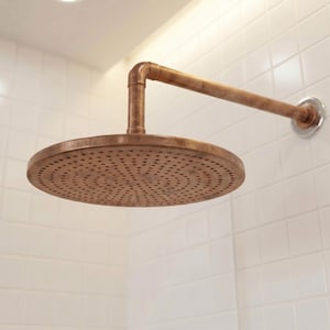 Op de afbeelding: Een ronde, koperkleurige douchekop met een verweerde afwerking en een bijpassende horizontale buisarm. De douchekop heeft een cirkelvormig patroon van kleine gaatjes voor de waterafvoer. Het armatuur is aan de muur bevestigd met een metalen plaat.