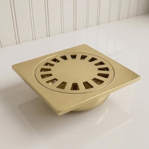 May include: A square, brushed brass drain cover with a circular, slotted center. The drain cover has a raised, rounded base and is designed for bathroom or shower use. The brass finish gives it a modern, minimalist aesthetic.