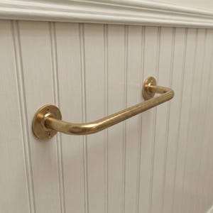 May include: A brass-colored, curved grab bar mounted on a white, vertical-paneled wall. The bar has circular mounting plates on each end, secured with visible screws. The design is simple and functional.