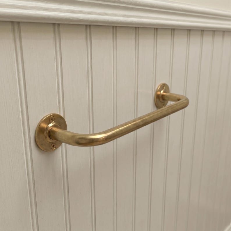 May include: A brass-colored, curved grab bar mounted on a white, vertical-paneled wall. The bar has circular mounting plates on each end, secured with visible screws. The design is simple and functional.