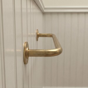 May include: A brass-colored, U-shaped grab bar mounted on a white wall. The bar has a smooth, curved design and is attached to the wall with circular mounting plates. The wall has vertical paneling.