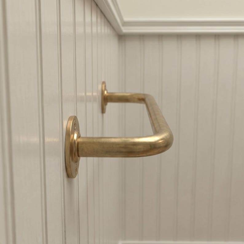 May include: A brass-colored, U-shaped grab bar mounted on a white wall. The bar has a smooth, curved design and is attached to the wall with circular mounting plates. The wall has vertical paneling.