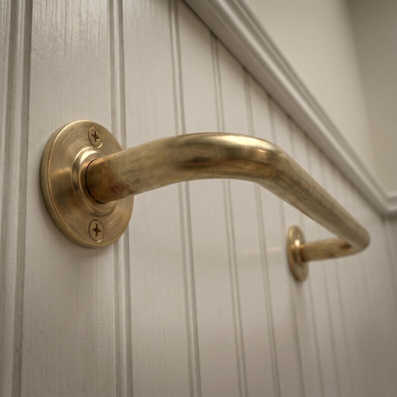 May include: A brass-colored grab bar mounted on a white, vertically paneled wall. The curved bar is attached with circular plates and screws. The design is simple and functional, suitable for bathroom or accessibility use.