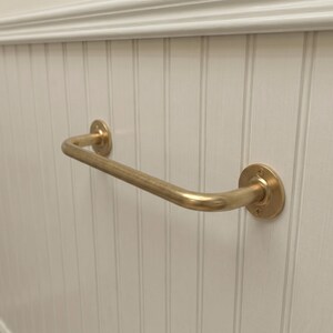 May include: A brass-colored, curved grab bar mounted on a white, vertical-paneled wall. The bar has a rounded shape and is attached to the wall with circular mounting plates. The wall has a decorative molding at the top.