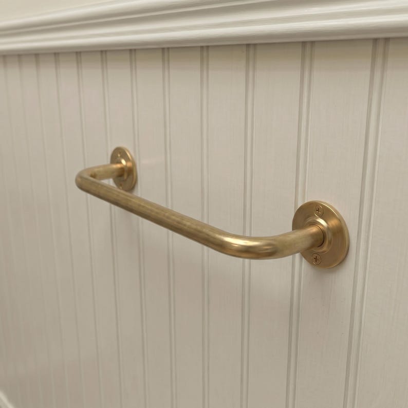 May include: A brass-colored, curved grab bar mounted on a white, vertical-paneled wall. The bar has a rounded shape and is attached to the wall with circular mounting plates. The wall has a decorative molding at the top.