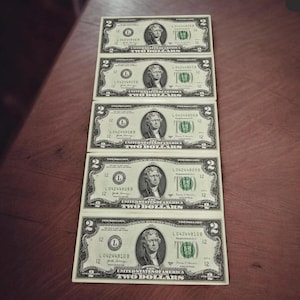 May include: Five United States two-dollar bills are arranged in a vertical stack on a wooden surface. Each bill features a portrait of Thomas Jefferson and the words "TWO DOLLARS". The bills are green and black.