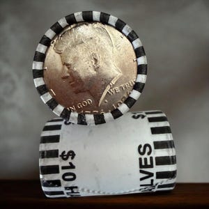 Puede incluir: Un rollo de monedas de medio dólar Kennedy con una envoltura a rayas blancas y negras. La moneda superior está fechada en 1971 y presenta un perfil de John F. Kennedy. La envoltura está etiquetada como "$10 HALVES".