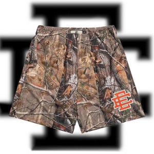 May include: Mesh shorts with a camouflage pattern in shades of brown, green, and orange. The shorts feature an orange and white logo on the lower right leg. The waistband is elastic. These shorts are designed for casual wear.