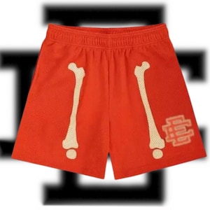 May include: Bright orange mesh shorts featuring bone designs on the front. The shorts have a cream-colored bone graphic on each leg and a cream-colored logo on the right leg. The shorts have an elastic waistband.