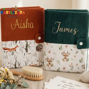 Può includere: Due album per bambini in velluto a coste, uno color ruggine con "Aisha" ricamato, l'altro verde con "James". Entrambi hanno una base in tessuto a tema bosco con un motivo di volpe, cervo e fiori. Sono visibili anche una spazzola di legno, fiori e scarpine.