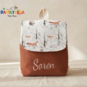 May include: A small, corduroy backpack with a flap featuring a woodland scene with foxes and trees. The lower portion is a rust color, and the flap has a white background with forest animals. The name "Saren" is embroidered on the front.