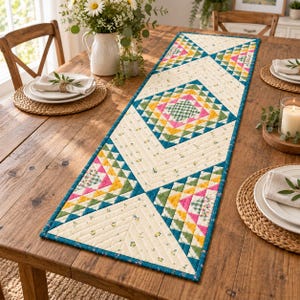 May include: A rectangular quilted table runner featuring a geometric pattern of triangles in shades of teal, yellow, pink, and green, interspersed with cream-colored fabric panels decorated with small floral prints and a dark teal fabric border.